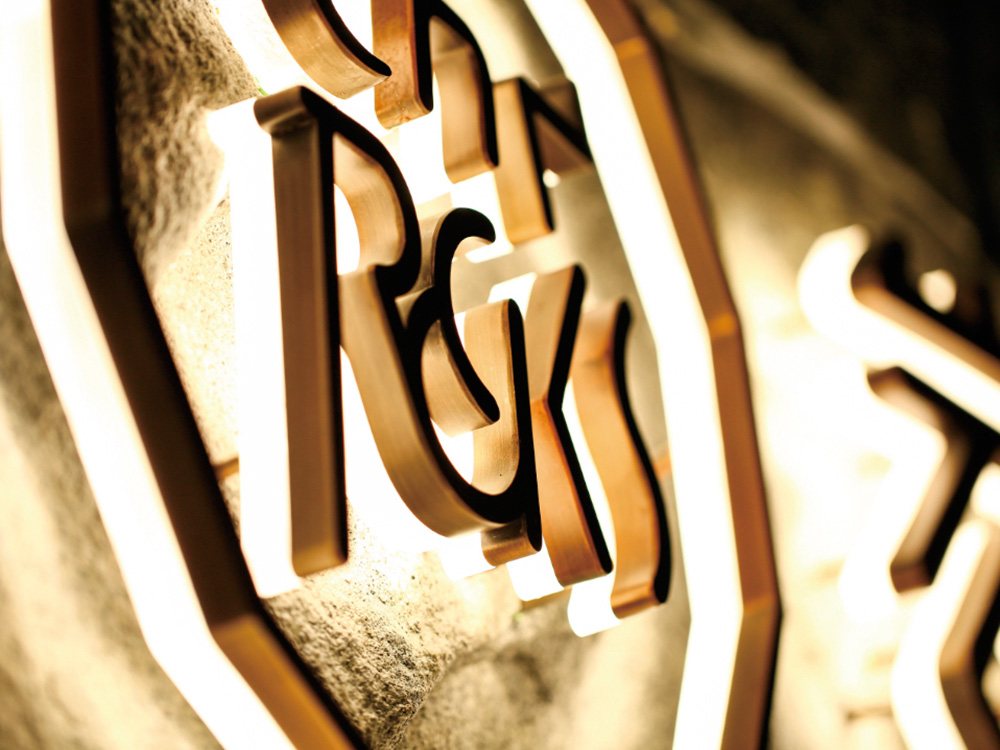 Close-up view of illuminated, raised lettering on a wall surface, possibly a logo or sign created by a signage company in Singapore. The letters appear to be made of a metallic material with a golden hue. The background is textured.