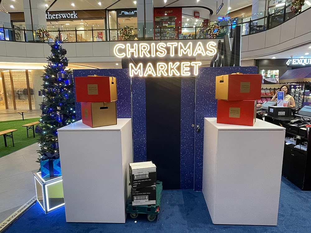 Indoor display with exhibition signage reading "Christmas Market," flanked by a decorated tree and stacked boxes on two pillars. Shopping mall setting.