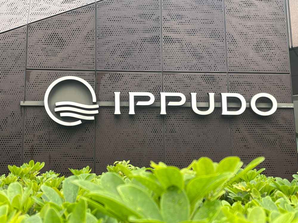 A metal wall displaying the "IPPUDO" logo and name, partially obscured by green shrubbery in the foreground, showcases the effectiveness of outdoor signs for business.