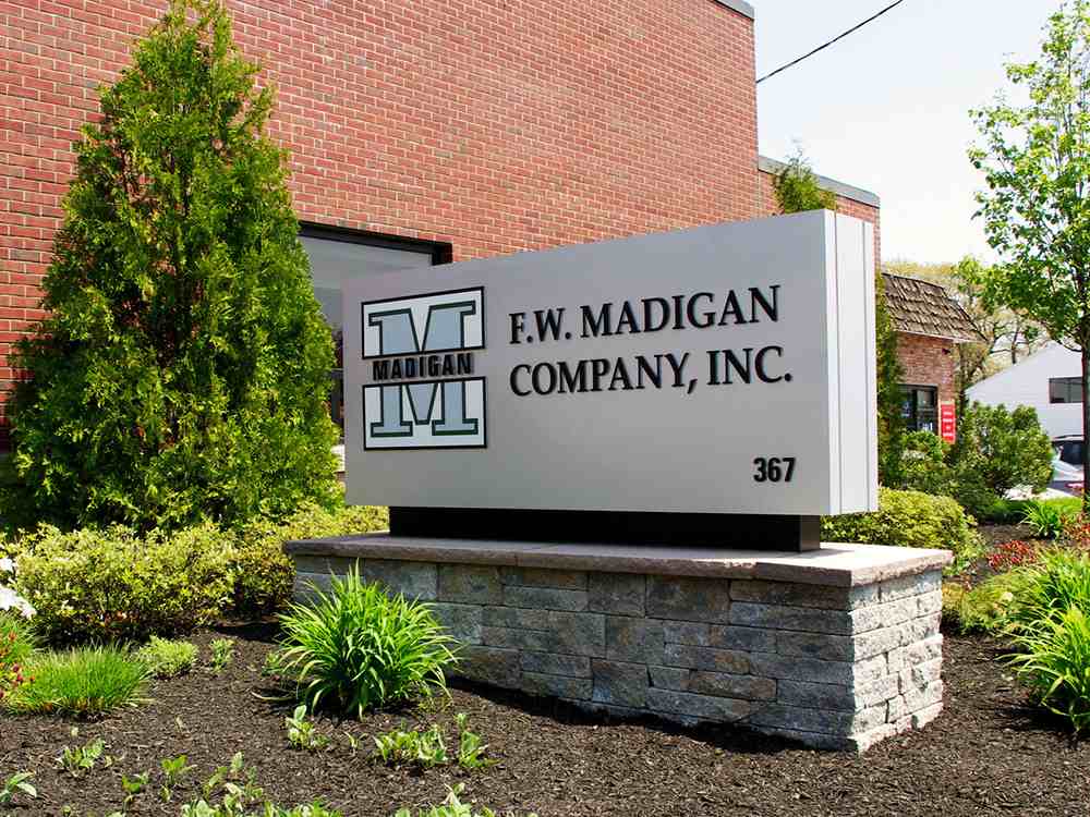 A sign outside a brick building reads "F.W. Madigan Company, Inc." with a large green "M" logo. This example of outdoor wayfinding signage is mounted on a stone base, surrounded by greenery.