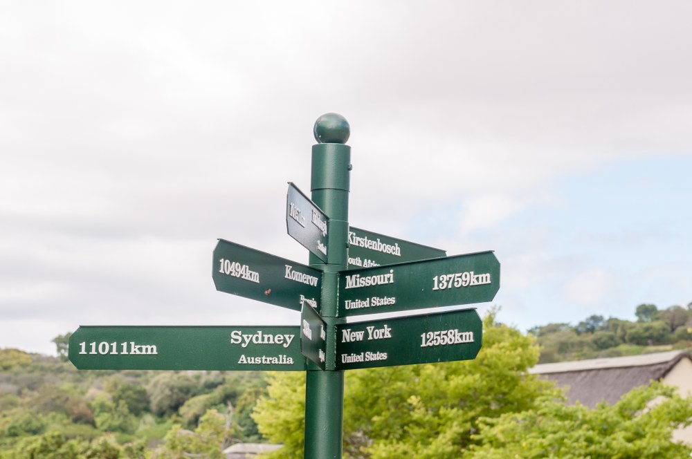 A multi-directional signpost, crafted by a signage company in Singapore, indicates distances to various locations including Sydney, New York, and Missouri against a backdrop of greenery and a cloudy sky.