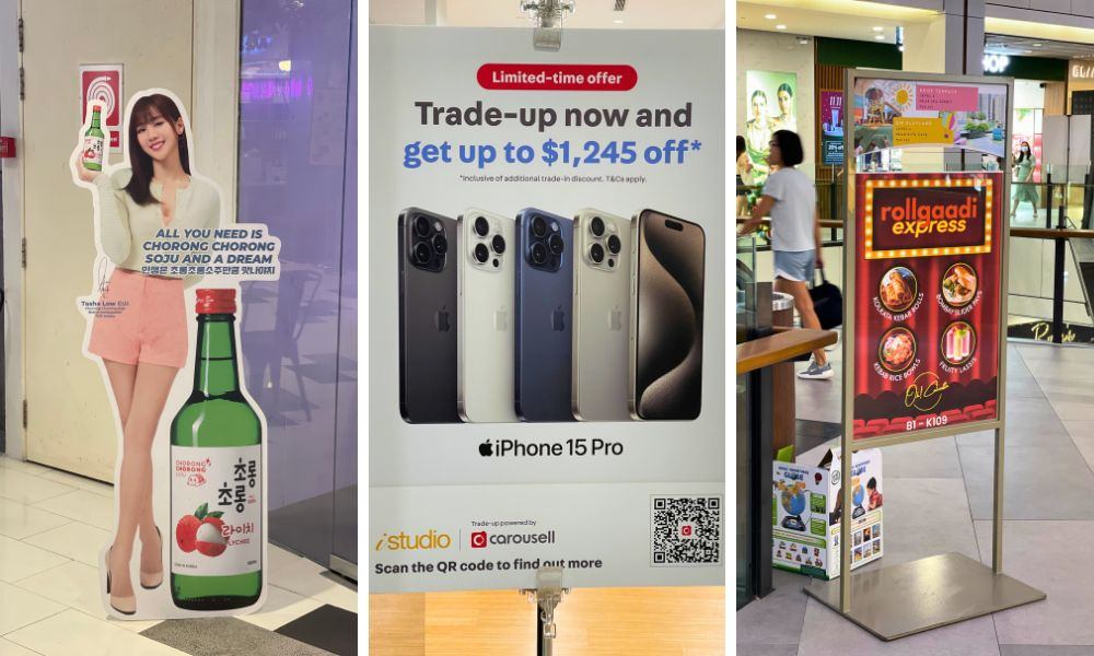 Three-panel image: an advertisement standee for a Korean drink with a model, an iPhone 15 Pro trade-up offer sign, and a rollgaadi express food stall banner. These vibrant displays highlight effective outdoor signage in Singapore's bustling shopping malls.