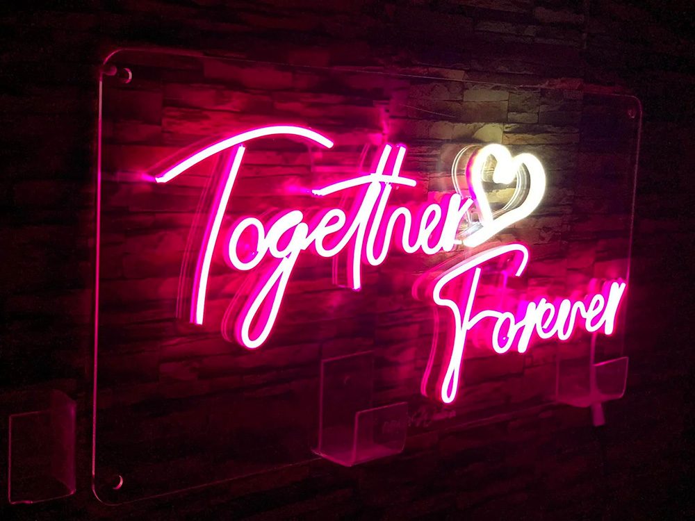 A neon sign crafted by a renowned signboard maker in Singapore, featuring the words "Together Forever" in pink cursive letters and a heart symbol, is beautifully mounted on a dark wall.