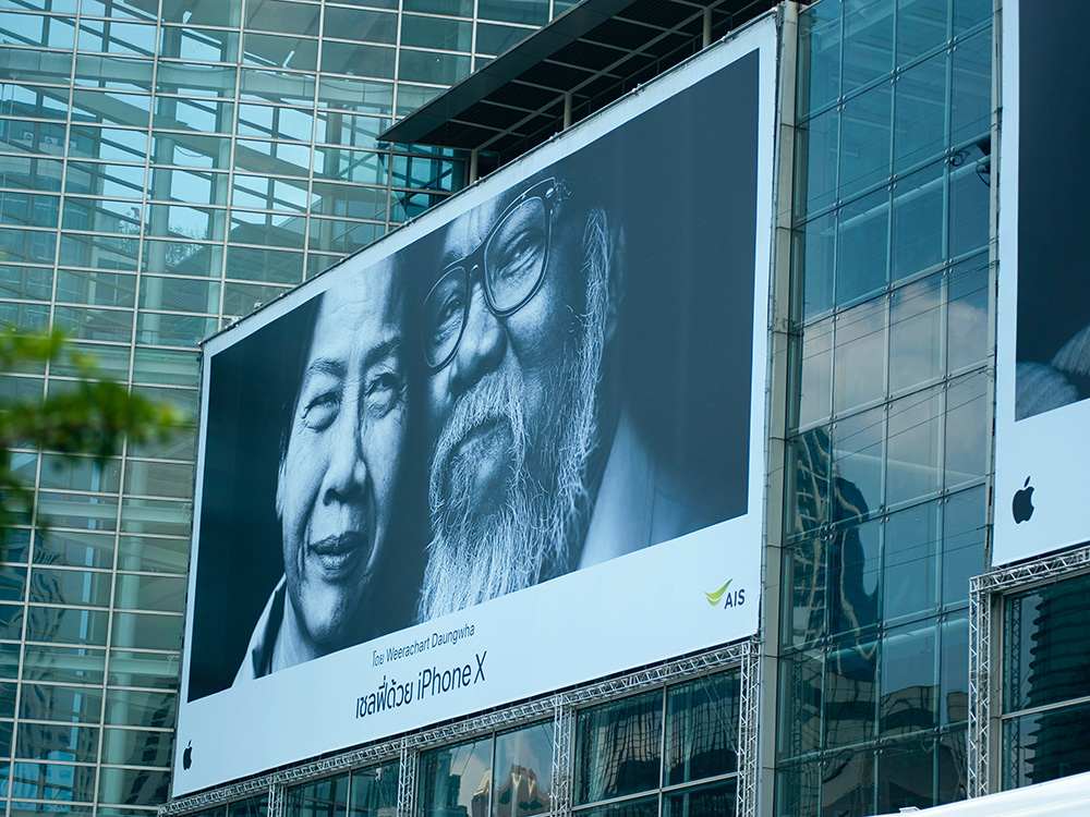 Large billboard advertisement for the iPhone X featuring a black-and-white photo of two smiling individuals, seamlessly integrated as outdoor signs for business on a glass building facade.